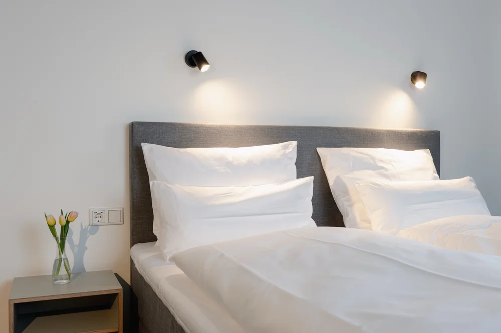 Modernes Hotelzimmer mit grauem Polsterbett, weißer Bettwäsche, zwei Wandlampen und einem Nachttisch mit frischen Tulpen in einer Glasvase.
