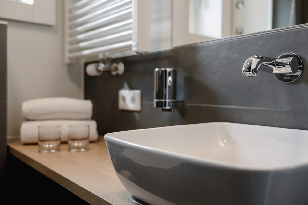 Detailaufnahme eines modernen Badezimmers mit eckigem Waschbecken, verchromtem Wandwasserhahn, Handtüchern und Gläsern auf einer Holzablage.