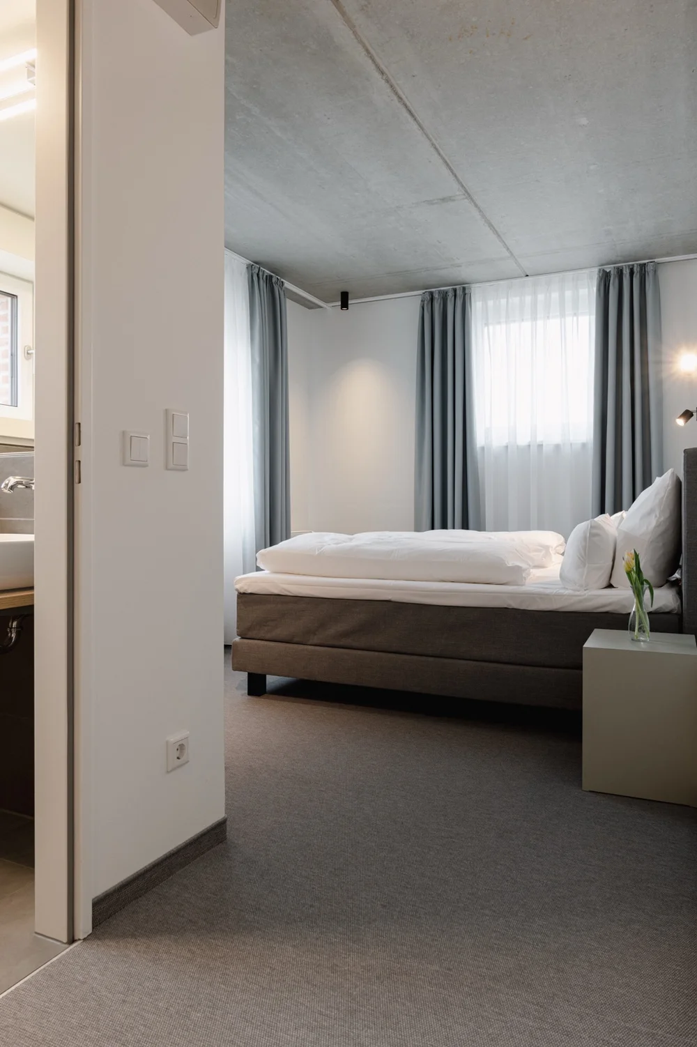 Helles Zimmer im Hotel baltica Hamburg Altona mit Boxspringbett, grauen Vorhängen, Sichtbetondecke und Blick ins angrenzende Badezimmer.