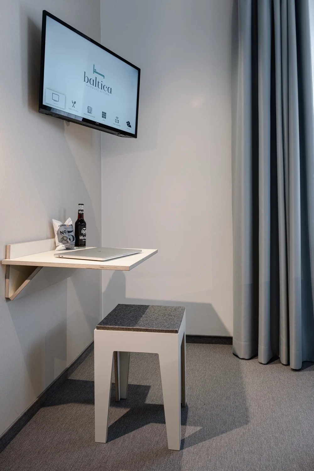 Kompakte Schreibtischecke im Hotel baltica Hamburg Altona mit Wandtisch, Hocker, Laptop, Getränk und TV über dem Arbeitsplatz.