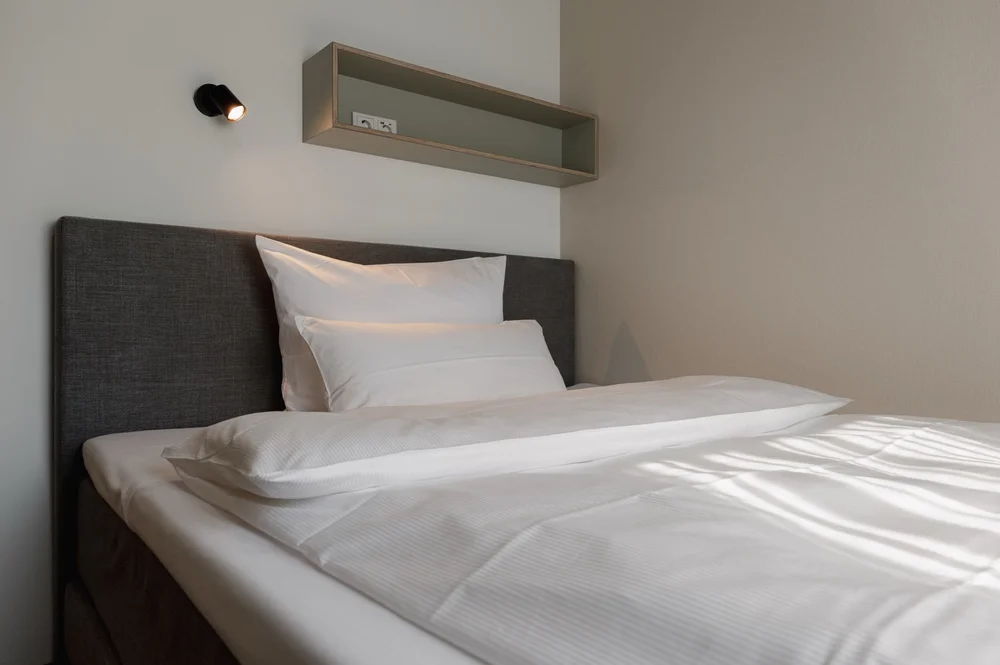 Minimalistisch eingerichtetes Einzelzimmer mit grauem Polsterbett, weißer Bettwäsche, Wandregal mit Steckdosen und moderner Leselampe.