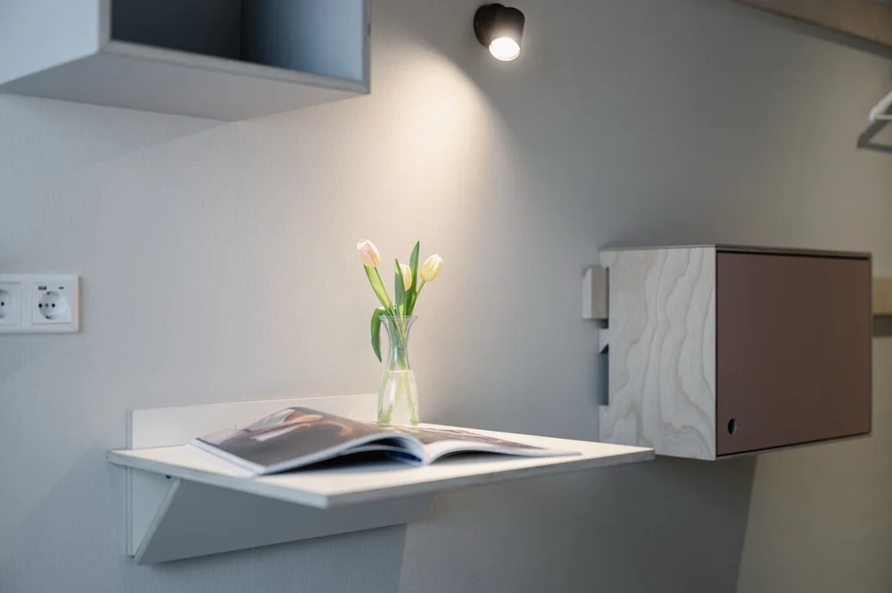 Detailaufnahme eines Hotelzimmers mit kleinem Wandtisch, geöffnetem Magazin, Vase mit Tulpen und minimalistischer Wandlampe