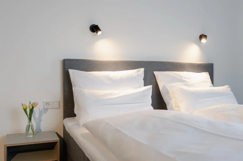 Modernes Hotelzimmer mit grauem Polsterbett, weißer Bettwäsche, zwei Wandlampen und einem Nachttisch mit frischen Tulpen in einer Glasvase.