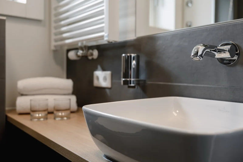 Modernes Badezimmer im Hotel baltica Hamburg Altona mit eckigem Waschbecken, verchromtem Wasserhahn, Handtüchern und Gläsern auf einer Holzablage