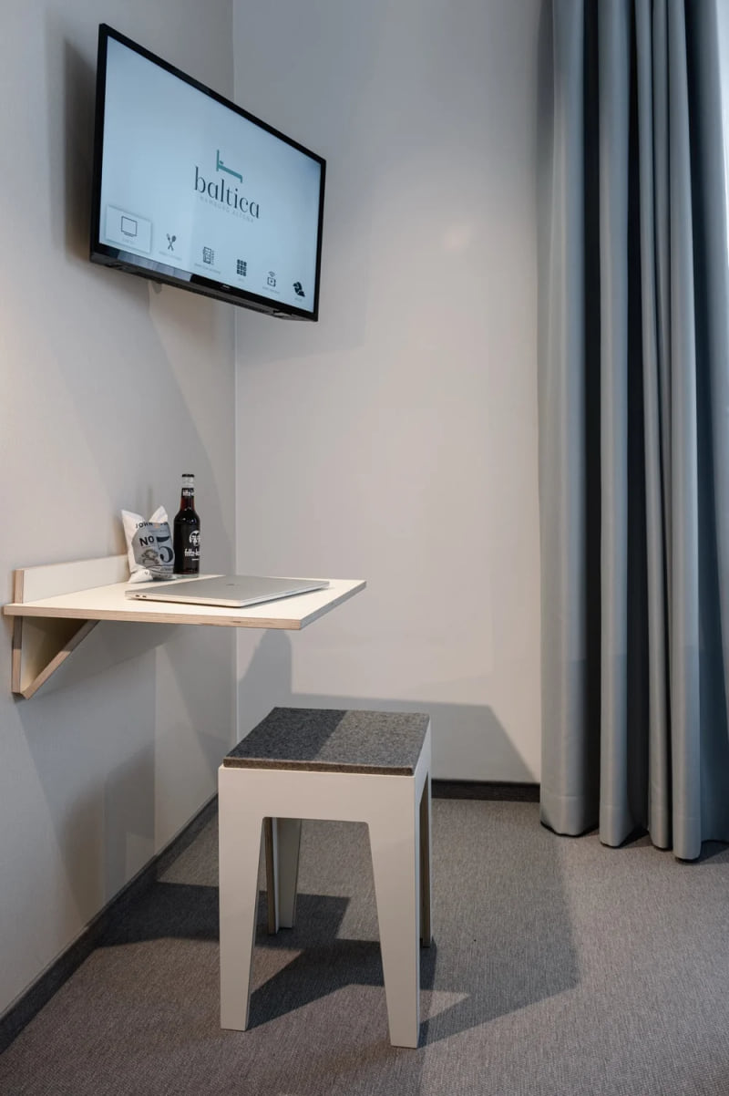 Kompakte Schreibtischecke im Hotel baltica Hamburg Altona mit Wandtisch, Hocker, Laptop, Getränk und TV über dem Arbeitsplatz.