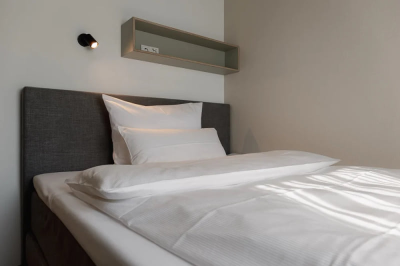 Minimalistisch eingerichtetes Einzelzimmer mit grauem Polsterbett, weißer Bettwäsche, Wandregal mit Steckdosen und moderner Leselampe.